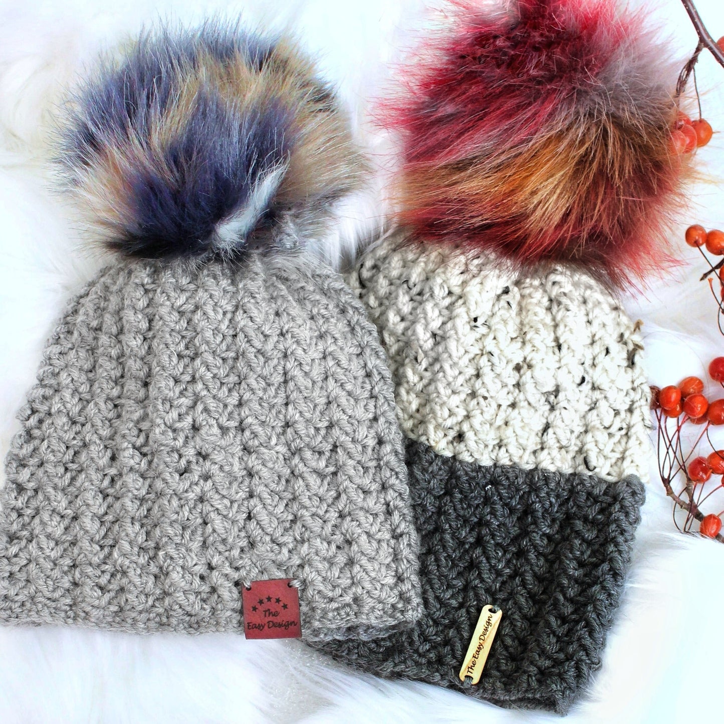 May include: Two crocheted hats with faux fur pom poms. The hat on the left is gray with a brown leather tag that says "The Easy Designs". The hat on the right is white, gray, and black with a brown leather tag that says "The Easy Designs".
