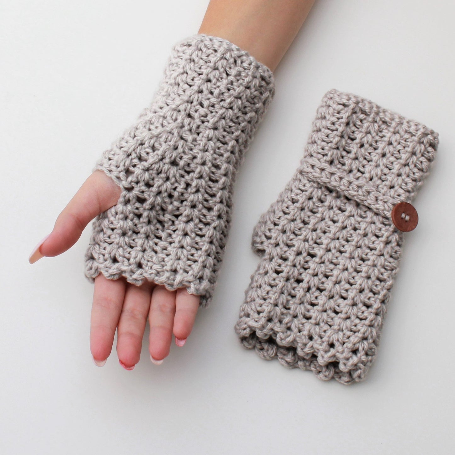 May include: A pair of beige crocheted fingerless gloves. One glove is on a hand, and the other glove has a brown button.