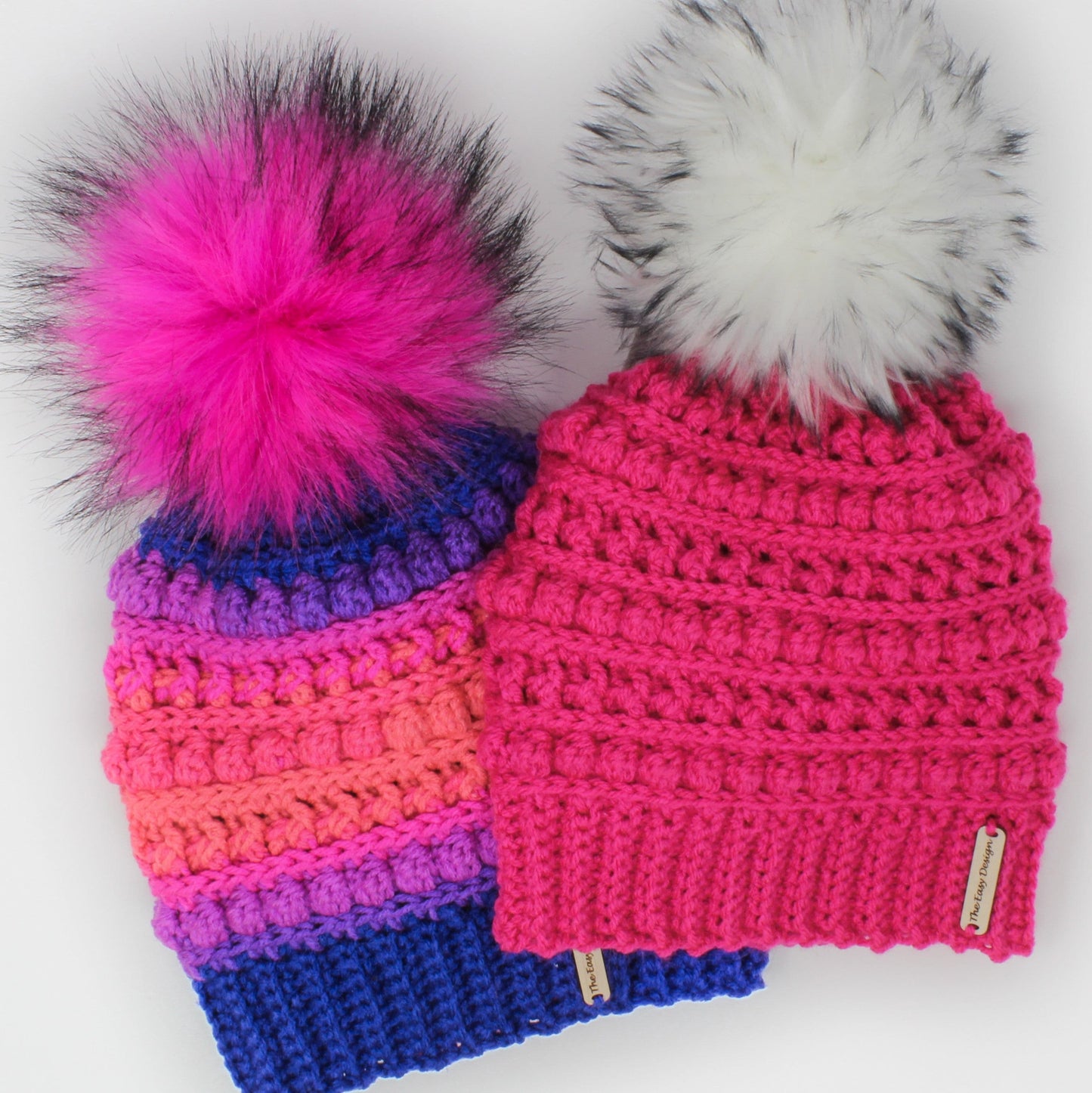 May include: Two crocheted hats with faux fur pom poms. One hat is pink and the other is a rainbow of colors. Both hats have a small leather tag with the text "The Yarn Designs".