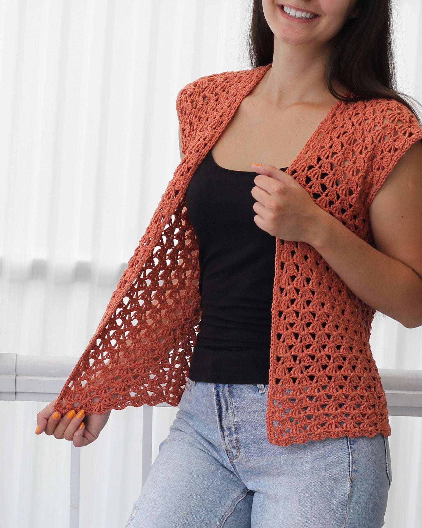 May include: A crocheted vest with a loose fit and a delicate openwork pattern. The vest is a light orange color and has a V-neckline. It is sleeveless and has a slightly cropped length.