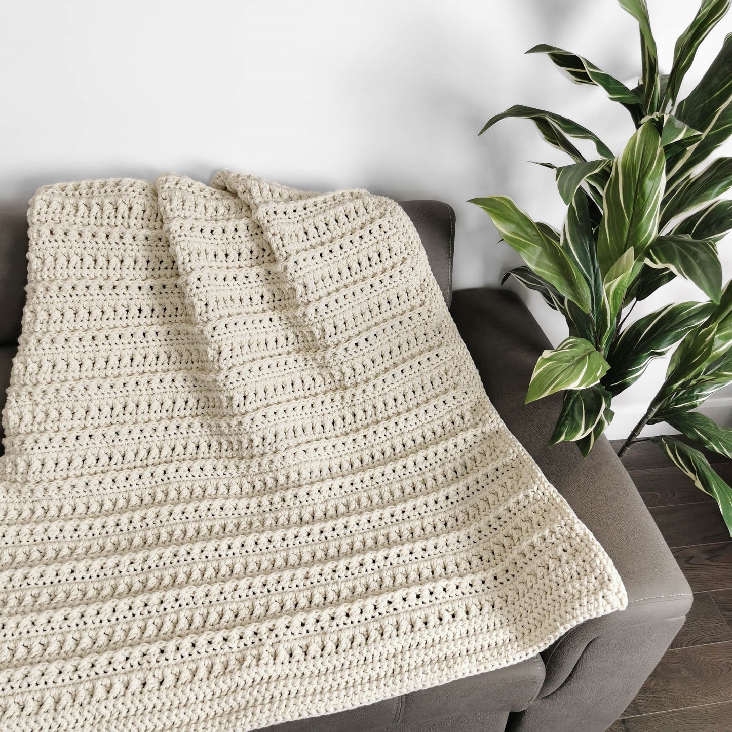 May include: A cream-colored crocheted throw blanket draped over a gray couch. The blanket has a textured stitch pattern and is made with a soft yarn.