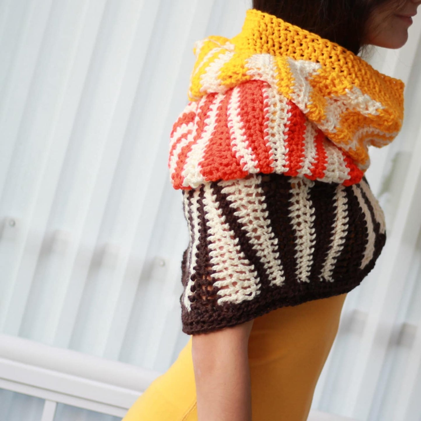 May include: A multi-colored crocheted cowl with stripes of orange, white, and brown. The cowl is worn by a person and is made of a soft, textured yarn.