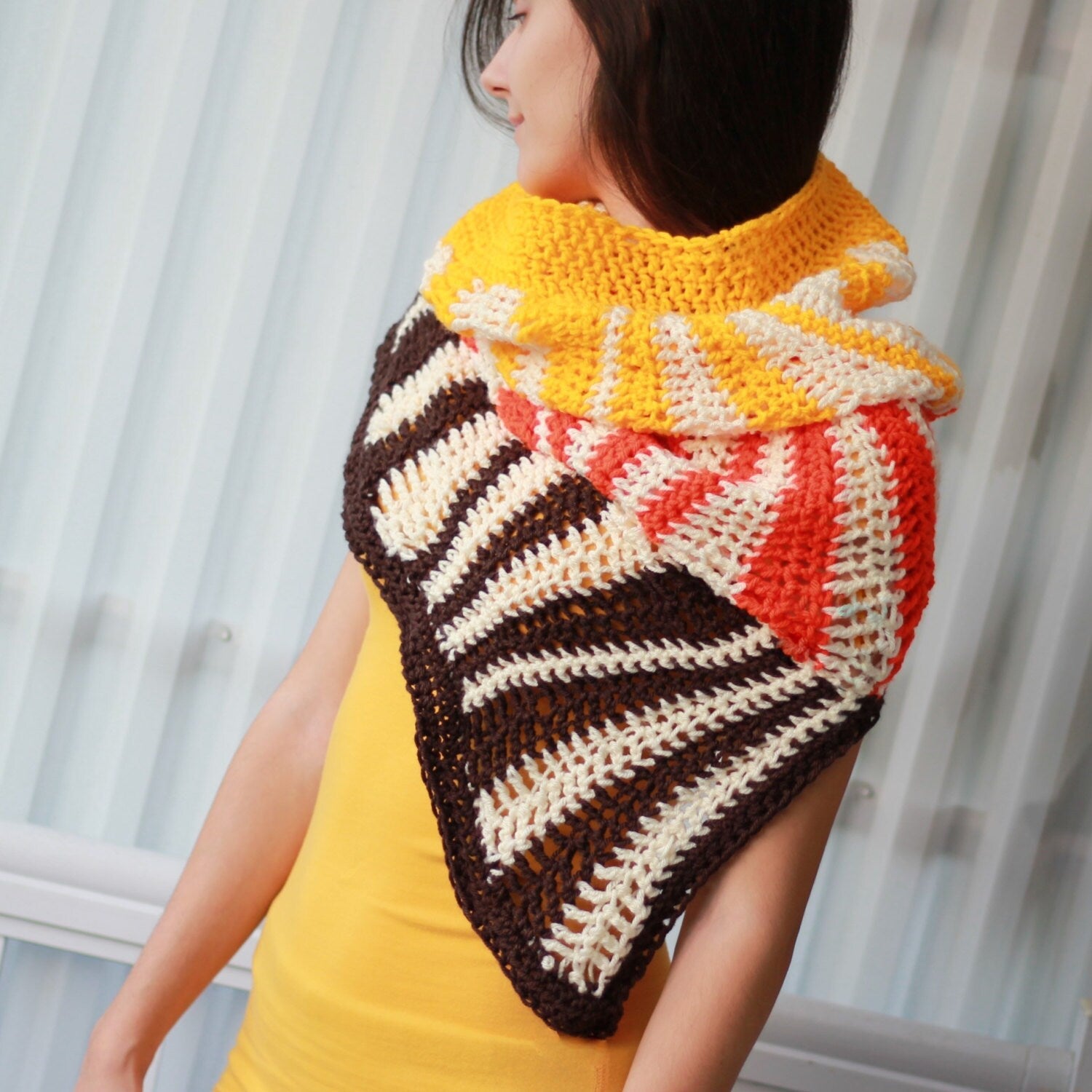 May include: A triangular crocheted shawl with brown, white, orange, and yellow stripes. The shawl is worn by a person with long dark hair.