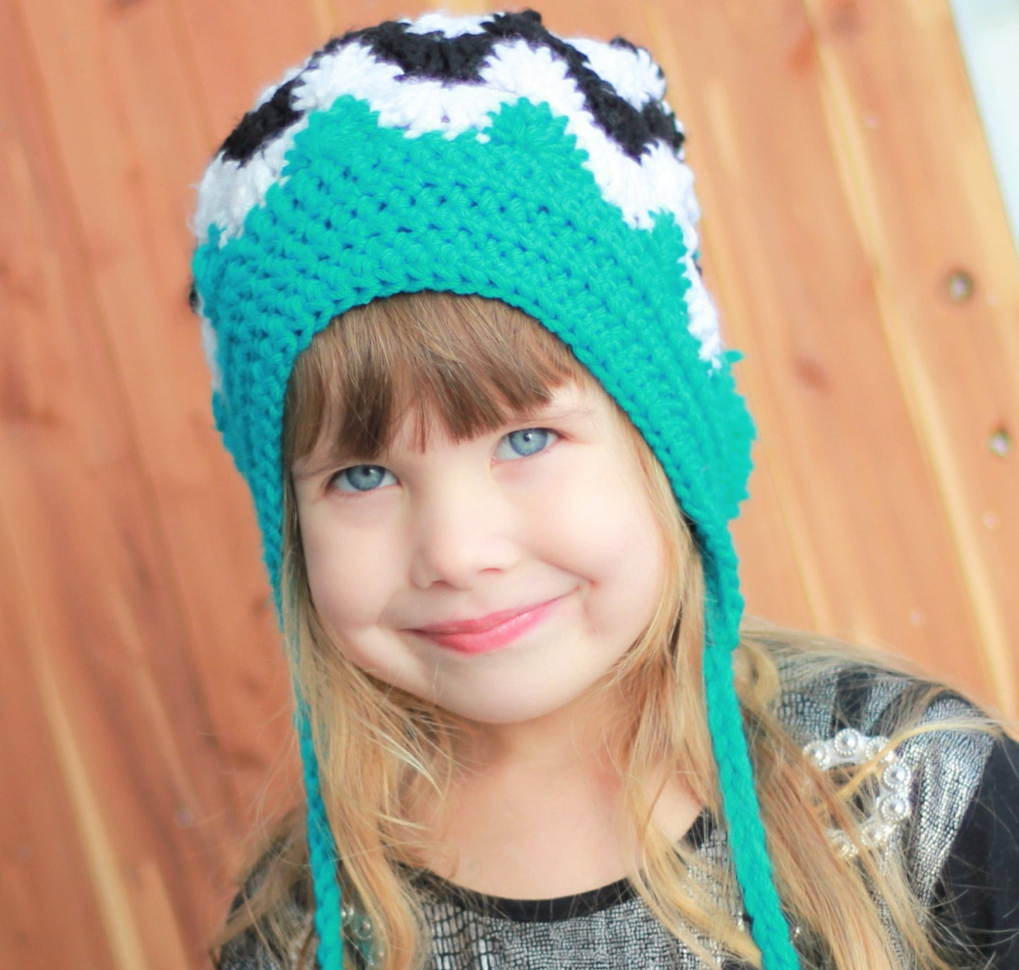 May include: A young girl wearing a teal and black crocheted hat with long ear flaps. The hat has a black and white geometric pattern.