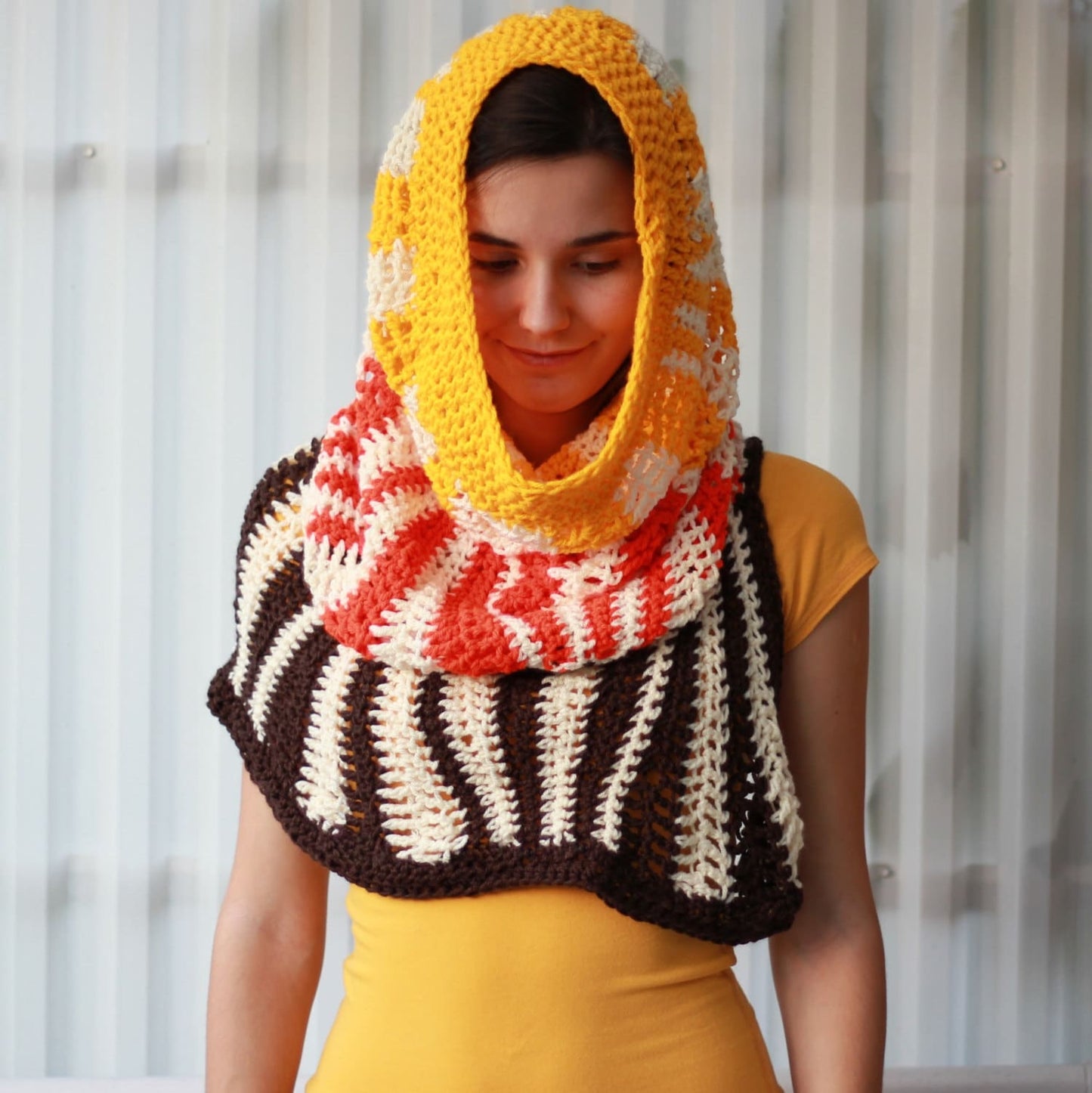 May include: A crocheted cowl with a hood in brown, white, orange, and yellow stripes. The cowl is worn by a person with brown hair.