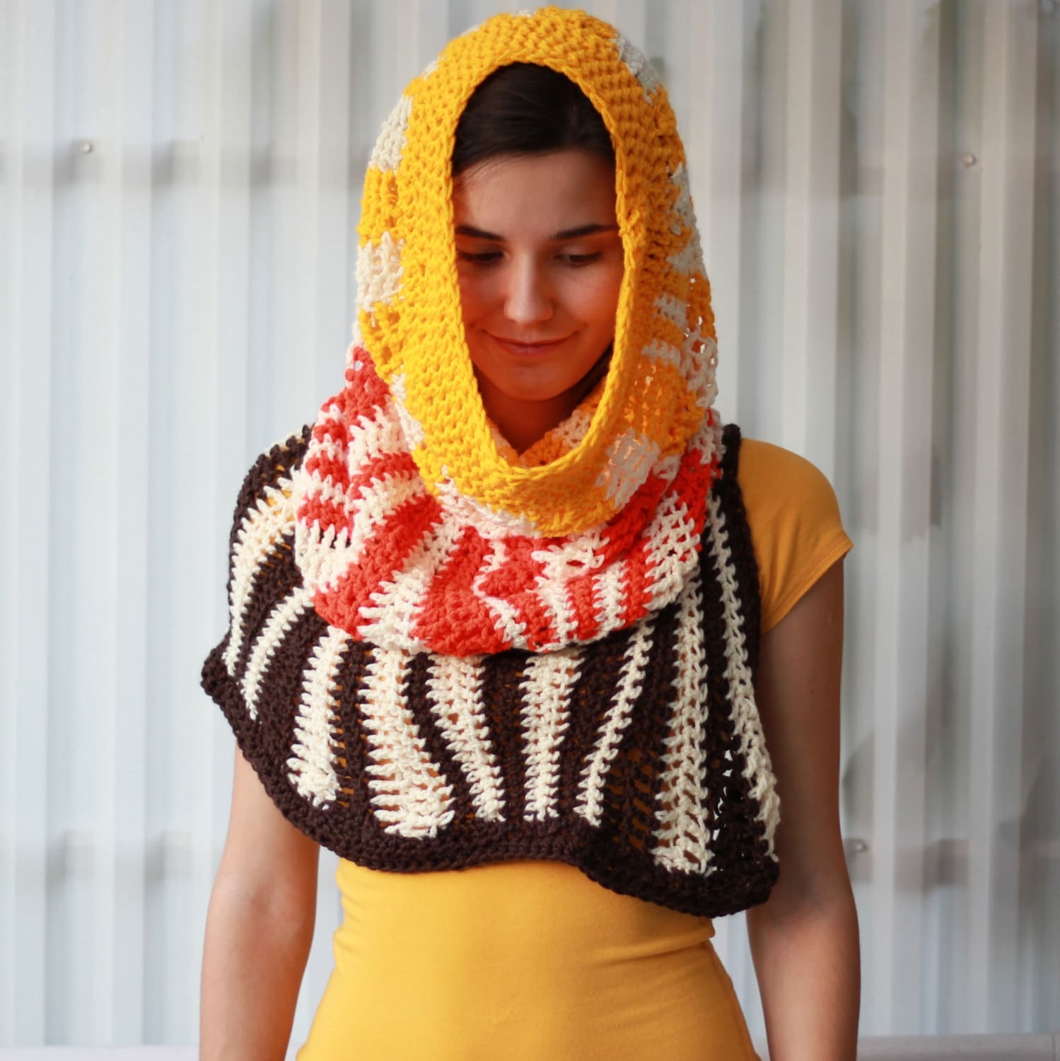 May include: A crocheted cowl with a hood in brown, white, orange, and yellow stripes. The cowl is worn by a person with brown hair.