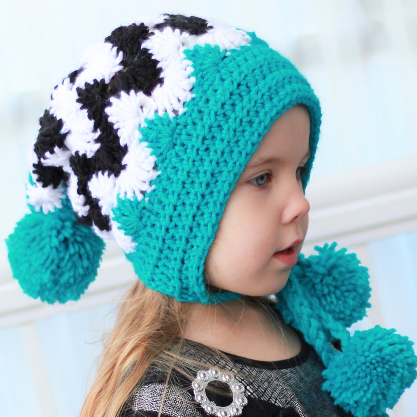 May include: A turquoise and black and white crocheted hat with pom poms on the ends of the ties. The hat has a pointed crown and ear flaps.