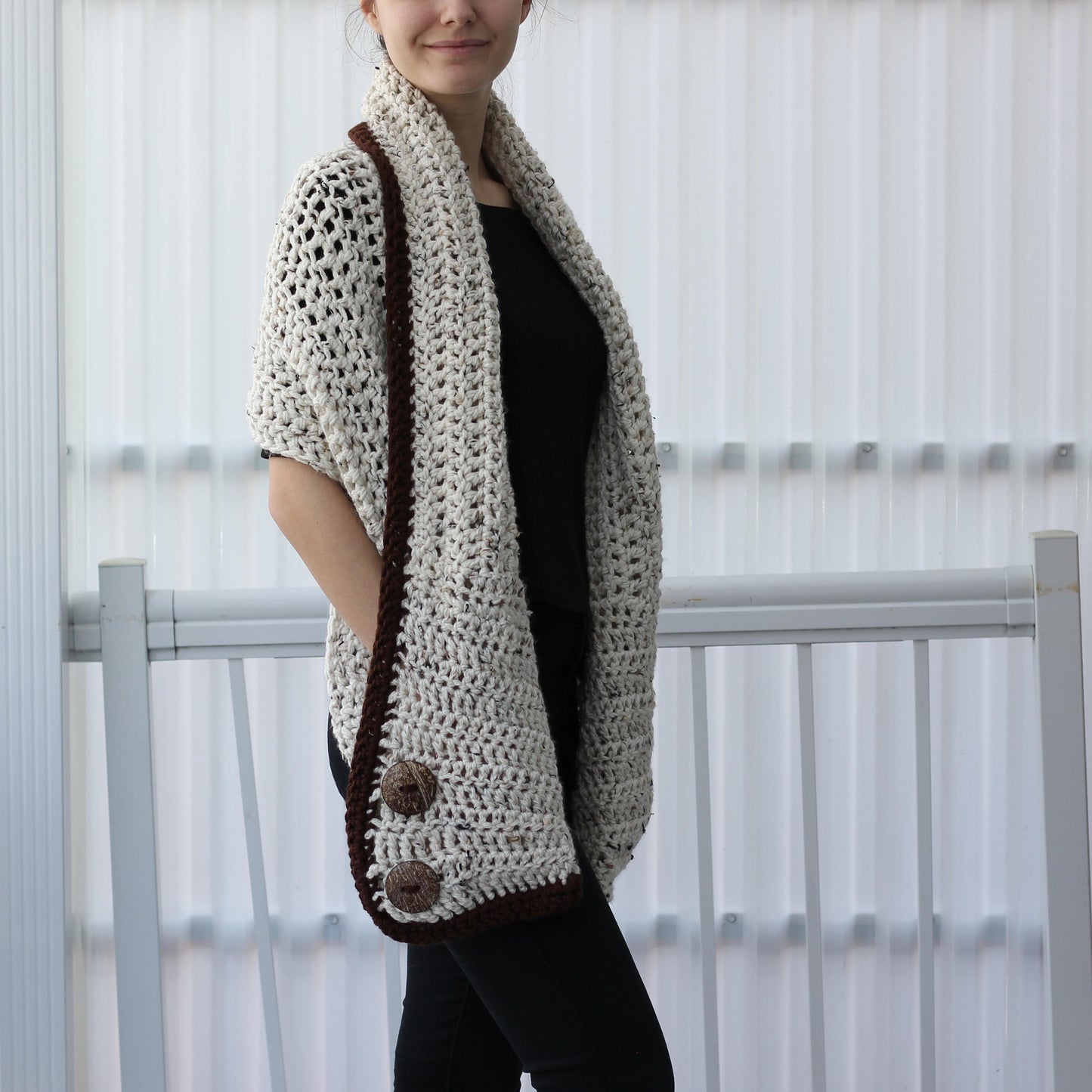 May include: A cream-colored, open-weave crochet shawl with a dark brown border and two large buttons. The shawl is draped over the shoulders, showcasing its textured design. The garment is a cozy accessory.