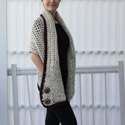 May include: A cream-colored, open-weave crochet shawl with a dark brown border and two large buttons. The shawl is draped over the shoulders, showcasing its textured design. The garment is a cozy accessory.