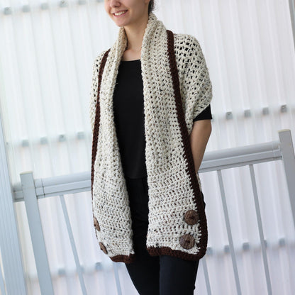 May include: A cream-colored, open-weave crochet shawl with a dark brown border and large wooden buttons. The shawl is draped over the shoulders, showcasing its textured design and contrasting colors. The buttons add a rustic touch to the handmade garment.