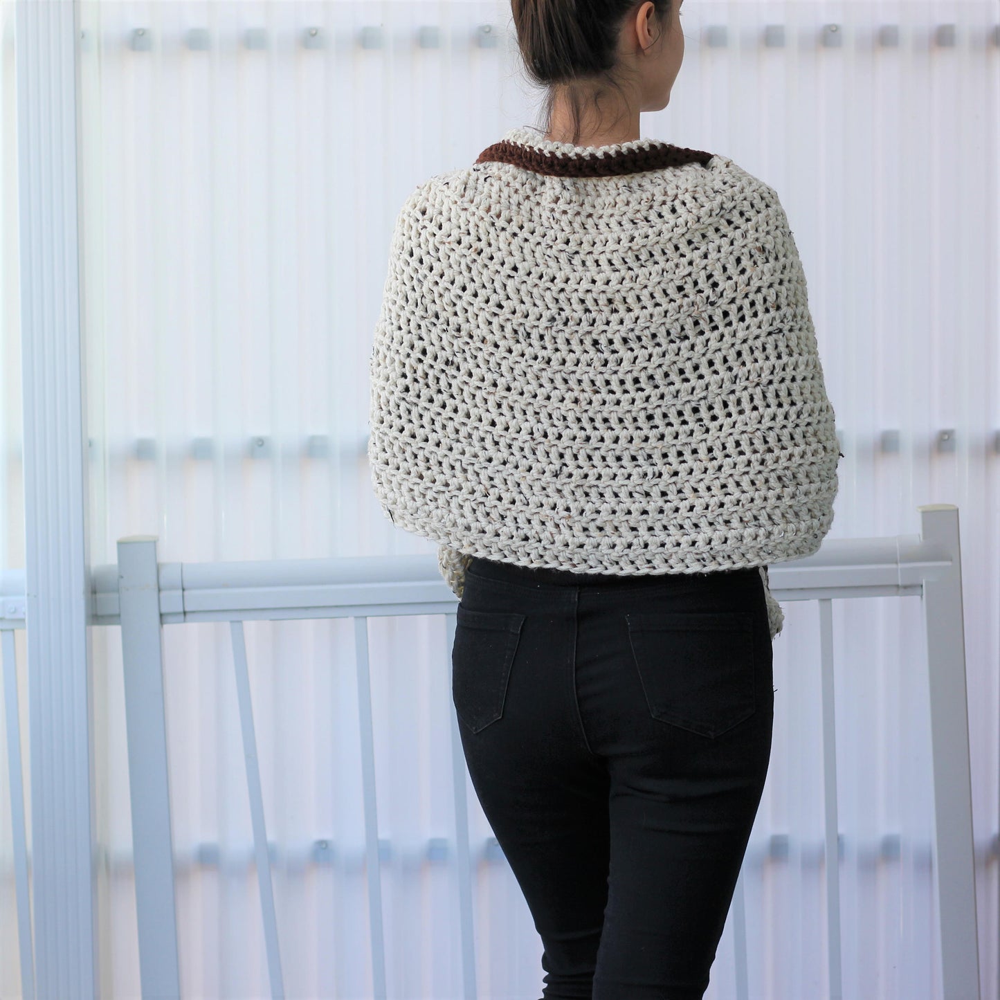 May include: A cream-colored, crocheted shawl with a brown collar. The open weave design of the shawl is visible. The person in the image is wearing black jeans. The shawl appears to be handmade.