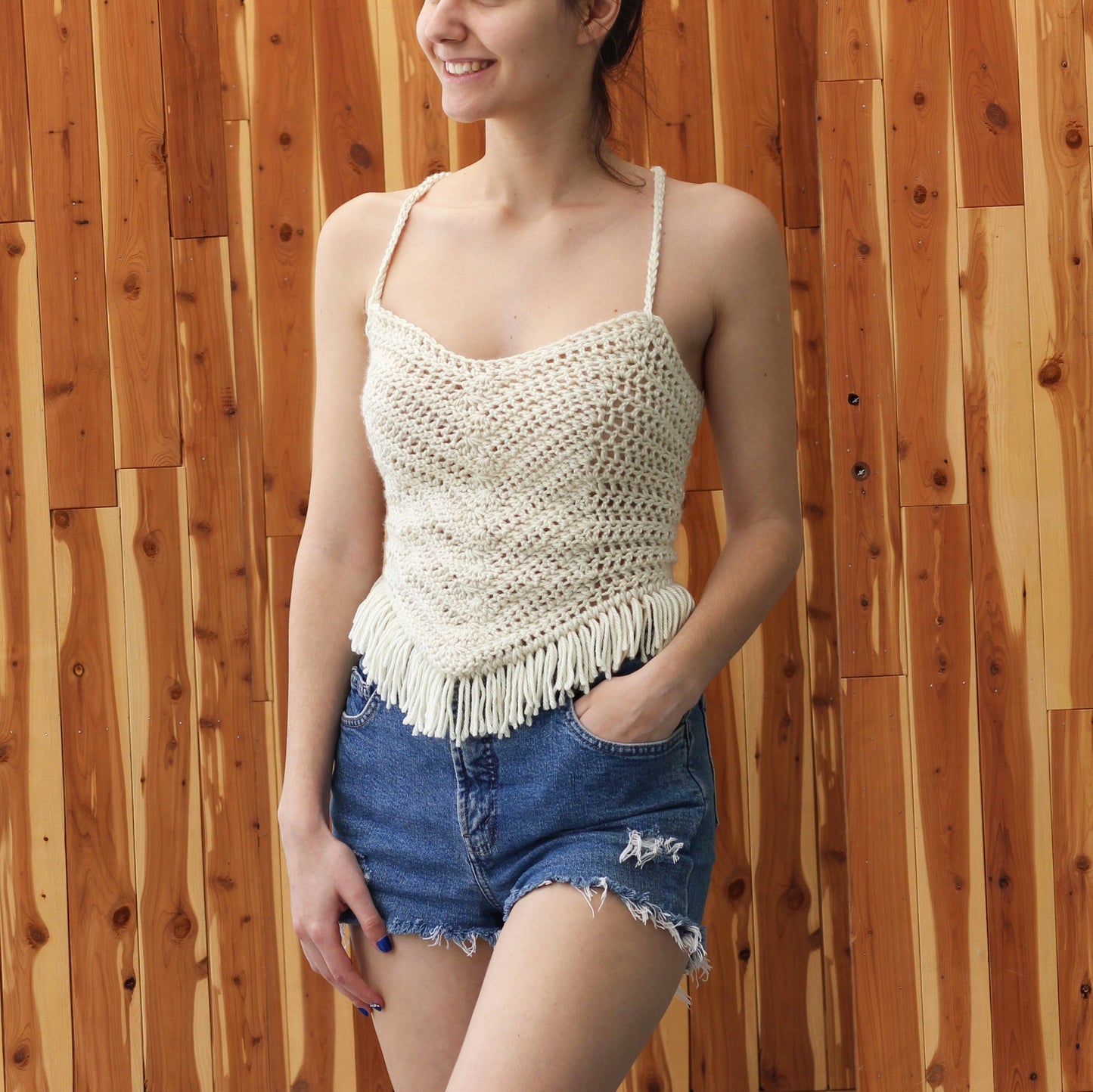 May include: A white crochet top with fringe detailing. The top has a scoop neck and thin straps. The model is wearing denim shorts.