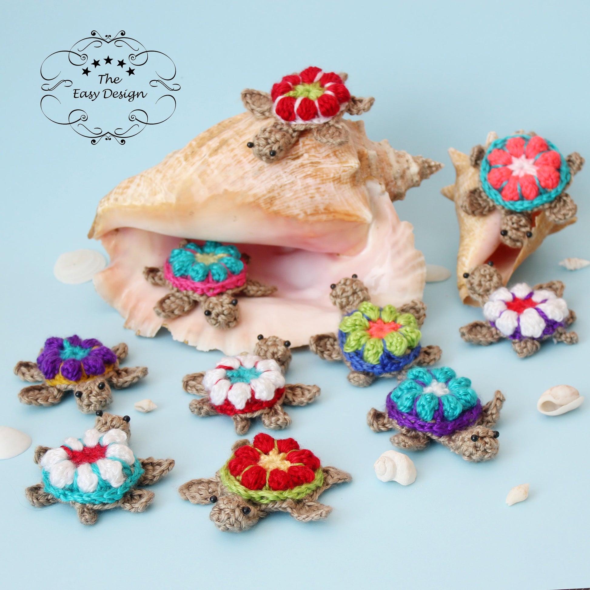 May include: A collection of nine crocheted sea turtles with colorful flower designs on their shells. The turtles are arranged around a large seashell on a light blue background. The text "The Easy Design" is visible in the top left corner of the image.