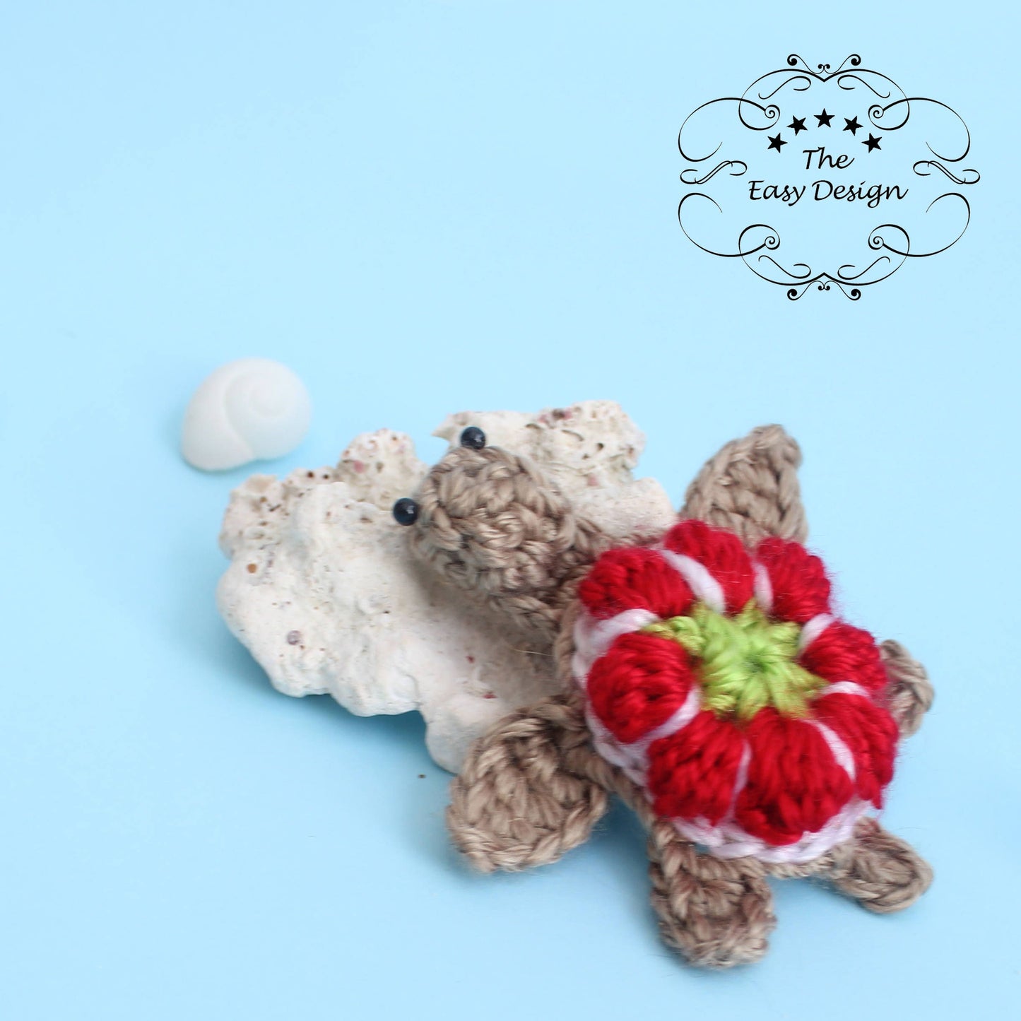 May include: A crocheted turtle toy with a red, white, and green shell. The turtle is sitting on a white seashell.