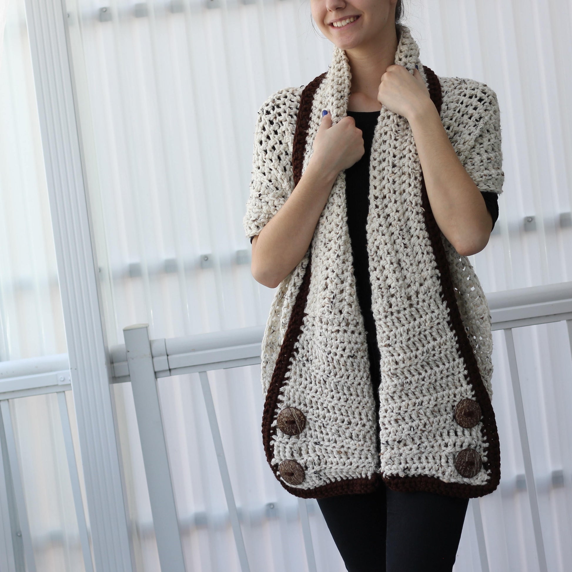 May include: Cream-colored crochet shawl with dark brown trim and large buttons. The open-front design features a textured stitch pattern and a wide collar. The shawl is a cozy and stylish accessory.