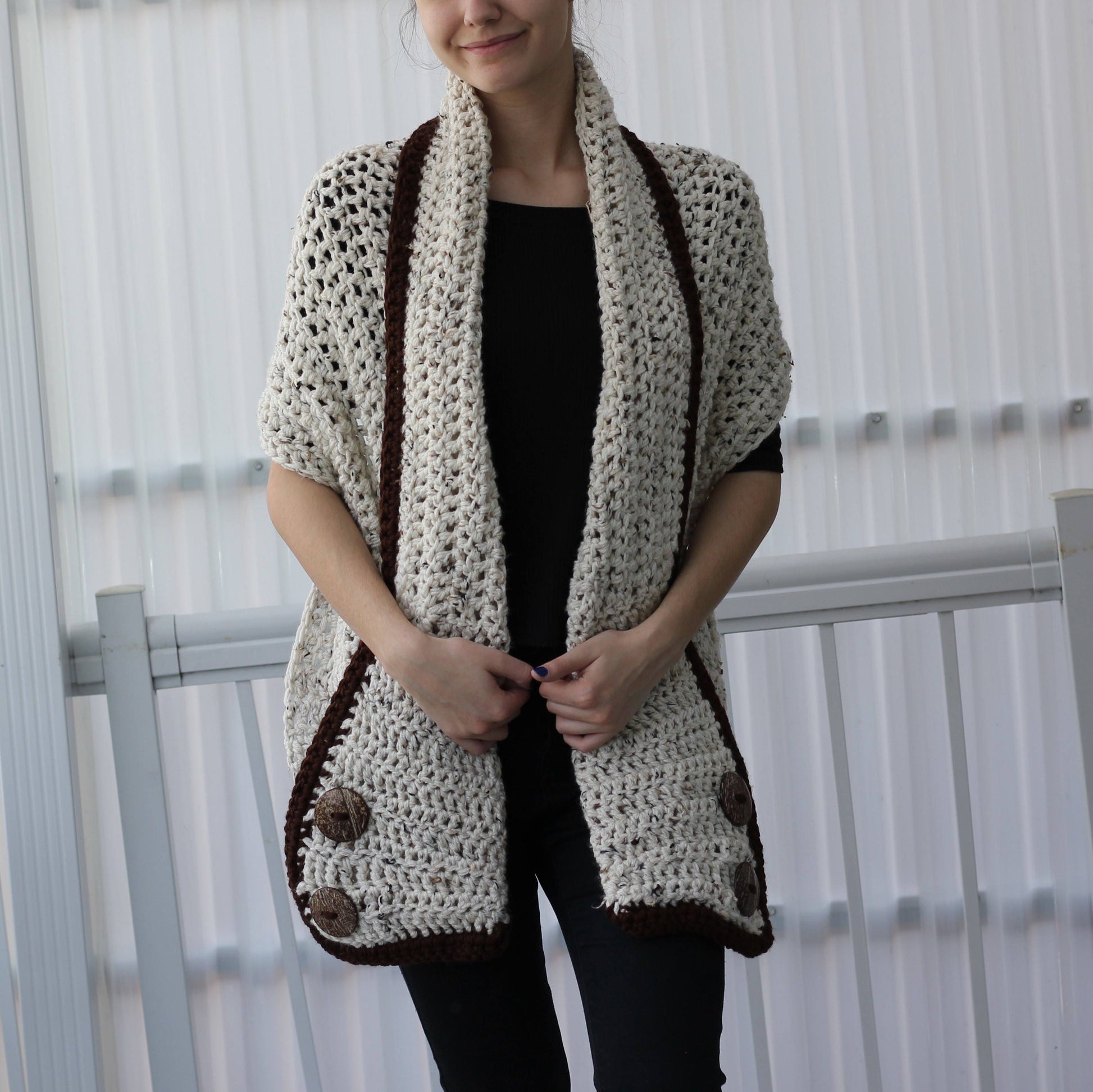 May include: Cream-colored crochet shawl with dark brown trim and large wooden buttons. The open-weave design features a shawl collar and short sleeves. This handmade garment is a cozy and stylish accessory.