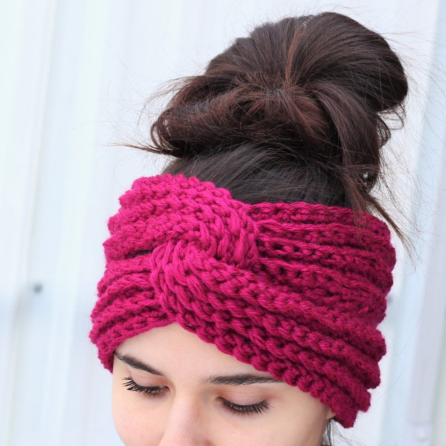 May include: A pink crocheted headband with a knotted design. The headband is worn by a person with brown hair.