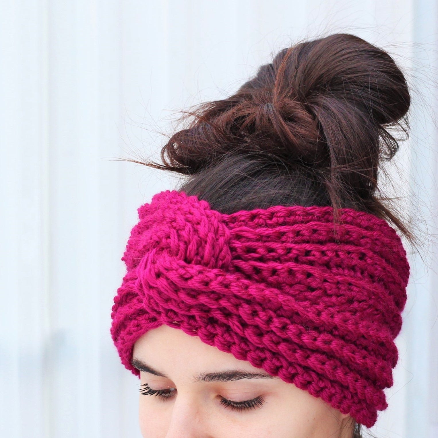 May include: A pink knitted headband with a knotted design. The headband is worn by a person with brown hair.