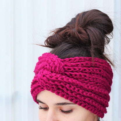 May include: A pink knitted headband with a knotted design. The headband is worn by a person with brown hair.