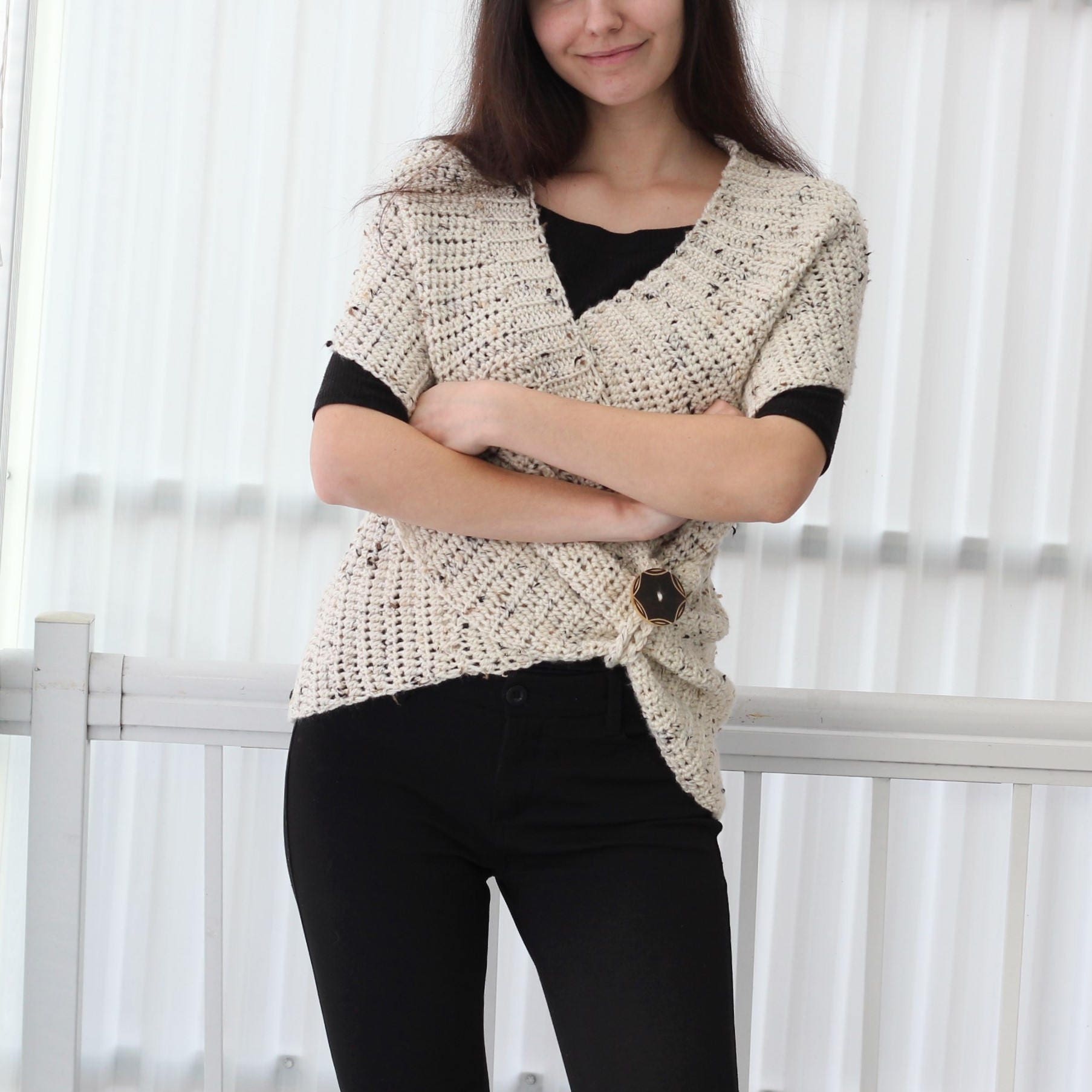 May include: A white crocheted wrap with a black button closure. The wrap has a speckled pattern and is worn over a black top and black pants.