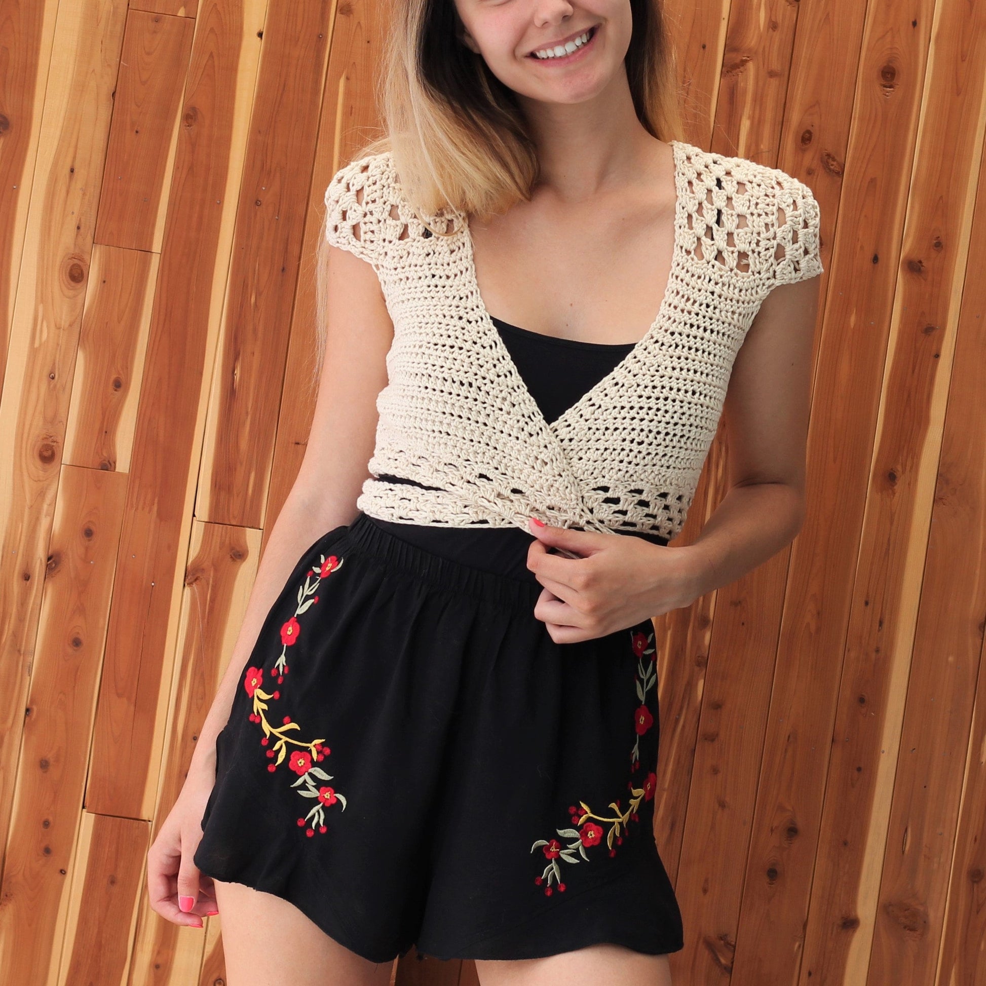 May include: A white crocheted wrap top with a V-neckline and short sleeves. The top is tied at the waist and worn over a black tank top. The model is wearing black shorts with red floral embroidery.