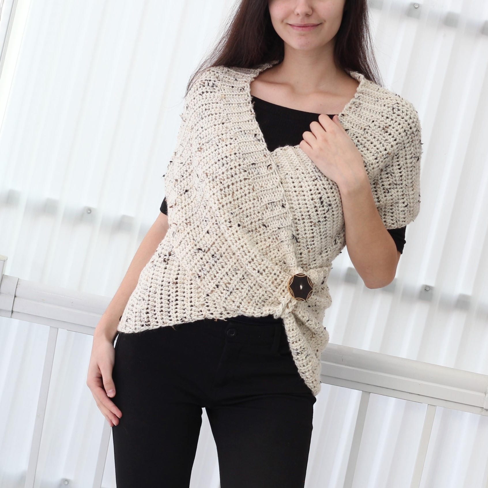May include: A white crocheted wrap with a speckled pattern and a black button closure. The wrap is worn over a black top and black pants.