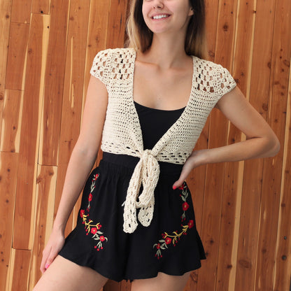 May include: A white crocheted crop top with a tie front and short sleeves. The top is worn over a black tank top and a black skirt with red floral embroidery.