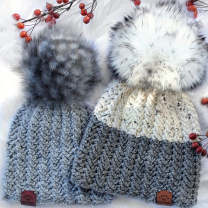 May include: Two crocheted hats with faux fur pom poms. The hats are gray and white with a chevron pattern. The pom poms are gray and white. The hats have a label that says "The Cozy Design".
