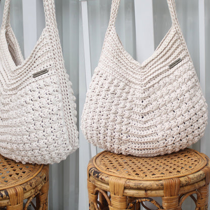May include: Two off-white, handmade crochet tote bags. The bags have a textured, bobble stitch pattern and long handles. Each bag has a small rectangular tag that reads "The Cozy Design". The bags are displayed on small, round wicker stools.