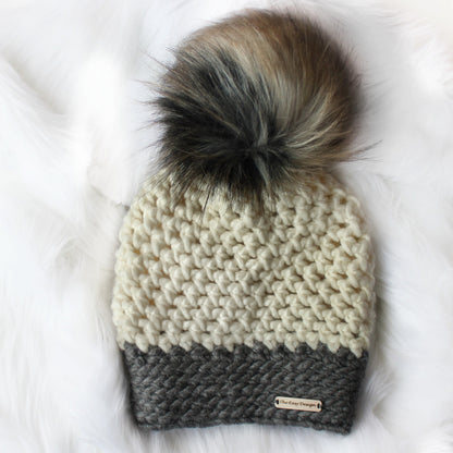 May include: A white and gray crocheted beanie with a faux fur pom pom. The beanie has a label that reads "The Easy Design".