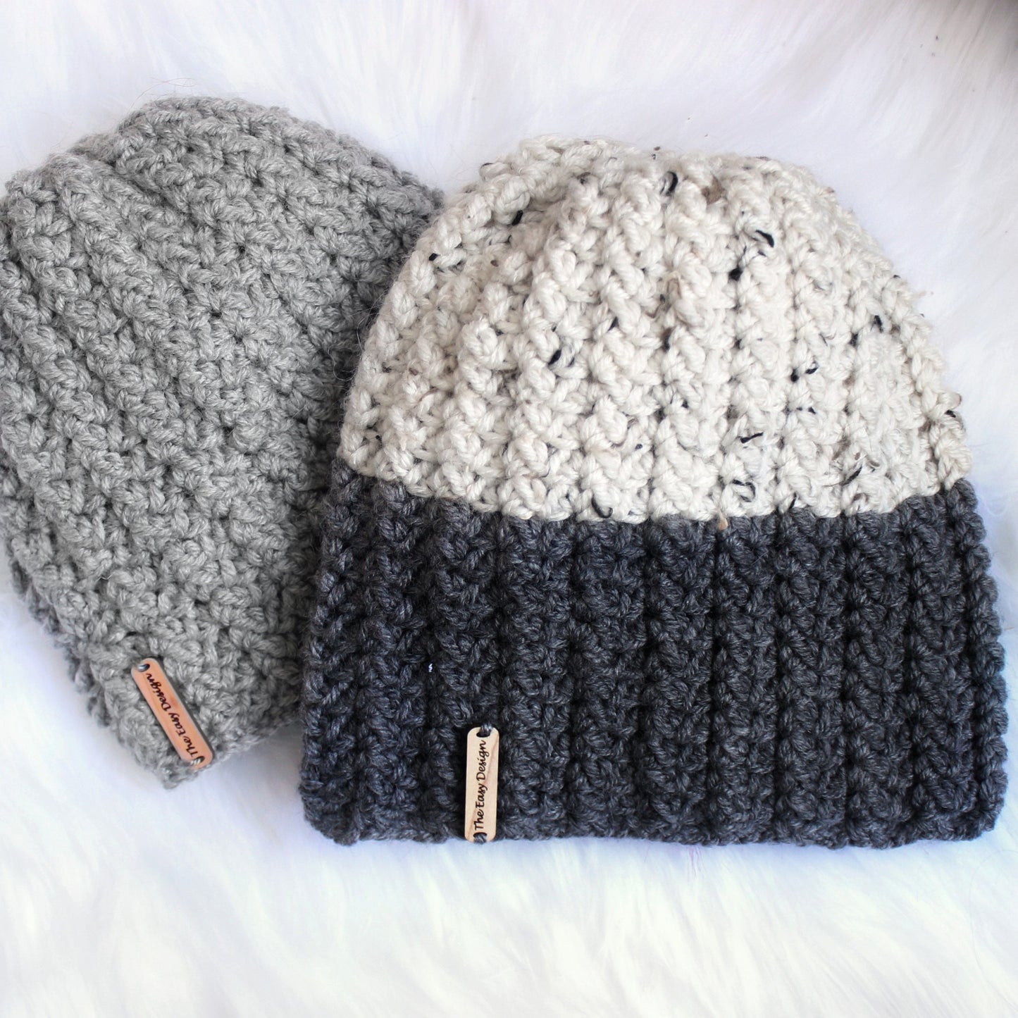 May include: Two crocheted hats, one gray and one with a white and gray top and a dark gray bottom. Both hats have a small brown tag with text that reads "The Easy Designer".