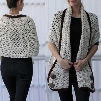 May include: Cream-colored crochet shawl with a brown border and large wooden buttons. The open-weave design is visible, and the shawl is draped over the shoulders. The garment is shown on a person wearing black pants and a black top. The shawl is a cozy accessory.