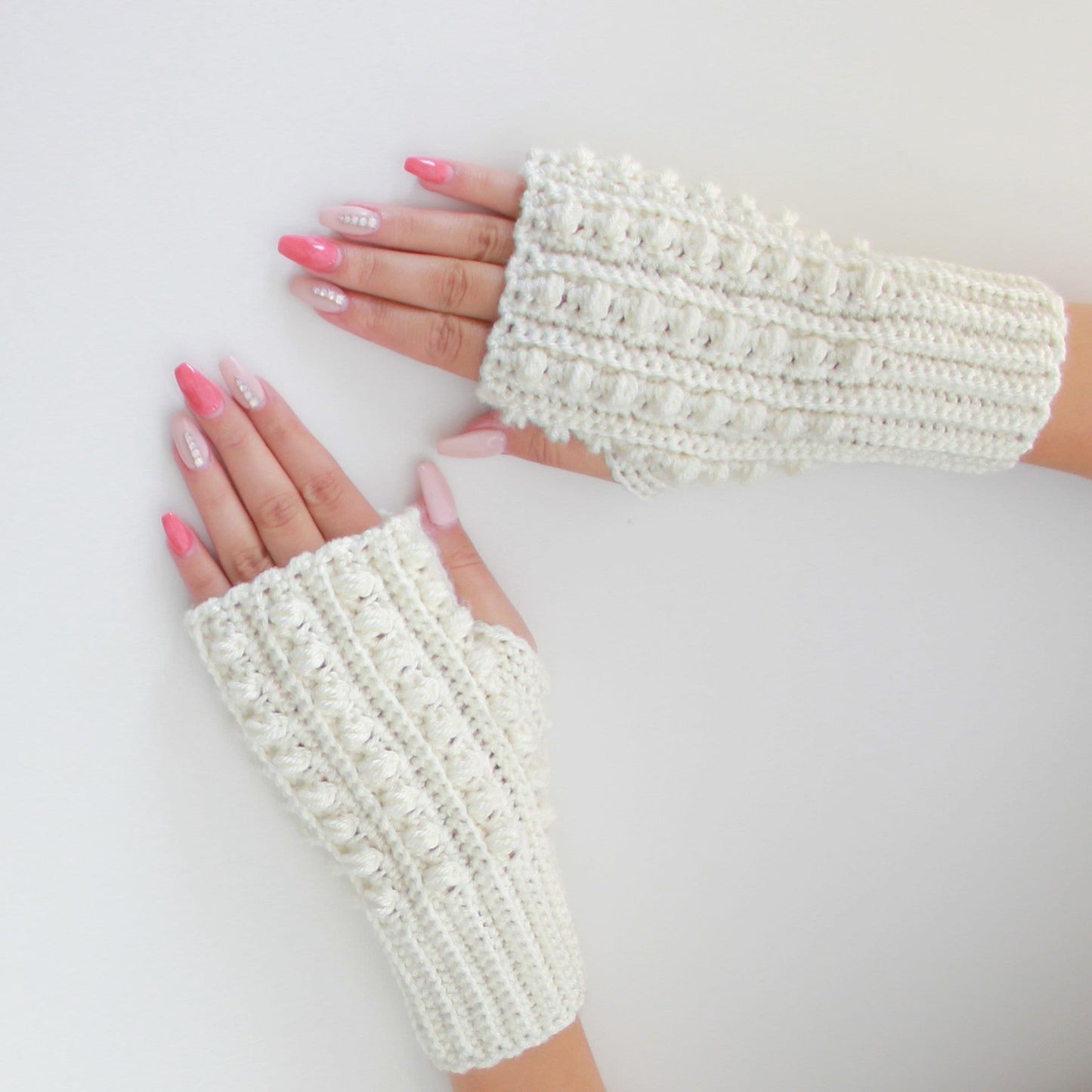 May include: A pair of white crocheted fingerless gloves with a textured design. The gloves are laid flat on a white surface.