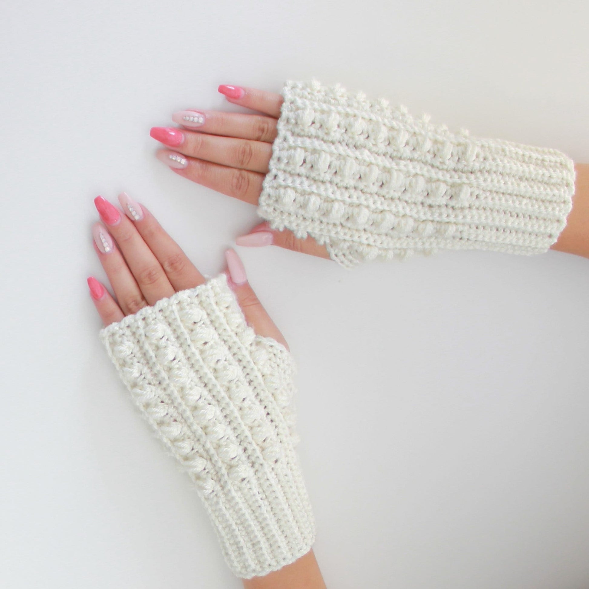 May include: A pair of white crocheted fingerless gloves with a textured design. The gloves are laid flat on a white surface.
