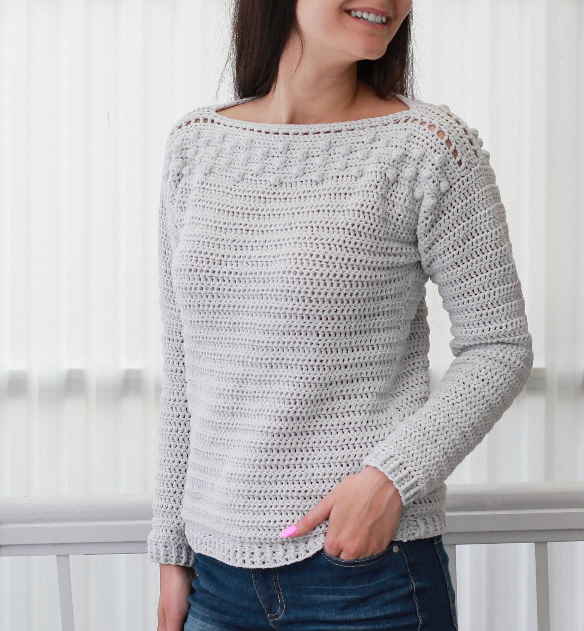 May include: A gray crocheted sweater with a boat neck and long sleeves. The sweater has a textured stitch pattern and a decorative trim around the neckline and shoulders.