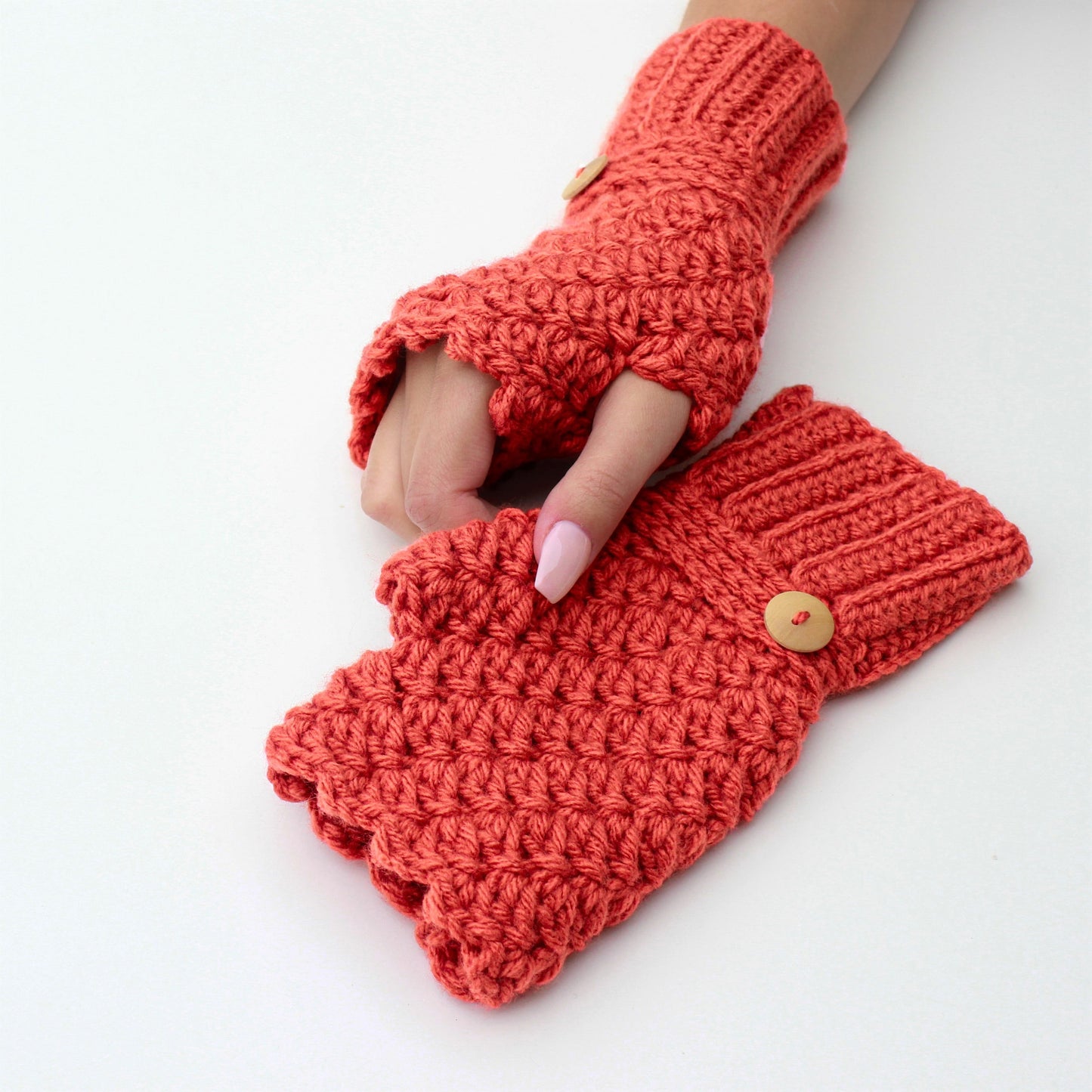 May include: A pair of coral-colored crocheted fingerless gloves with wooden buttons. The gloves are made with a textured stitch and have a fold-over cuff.