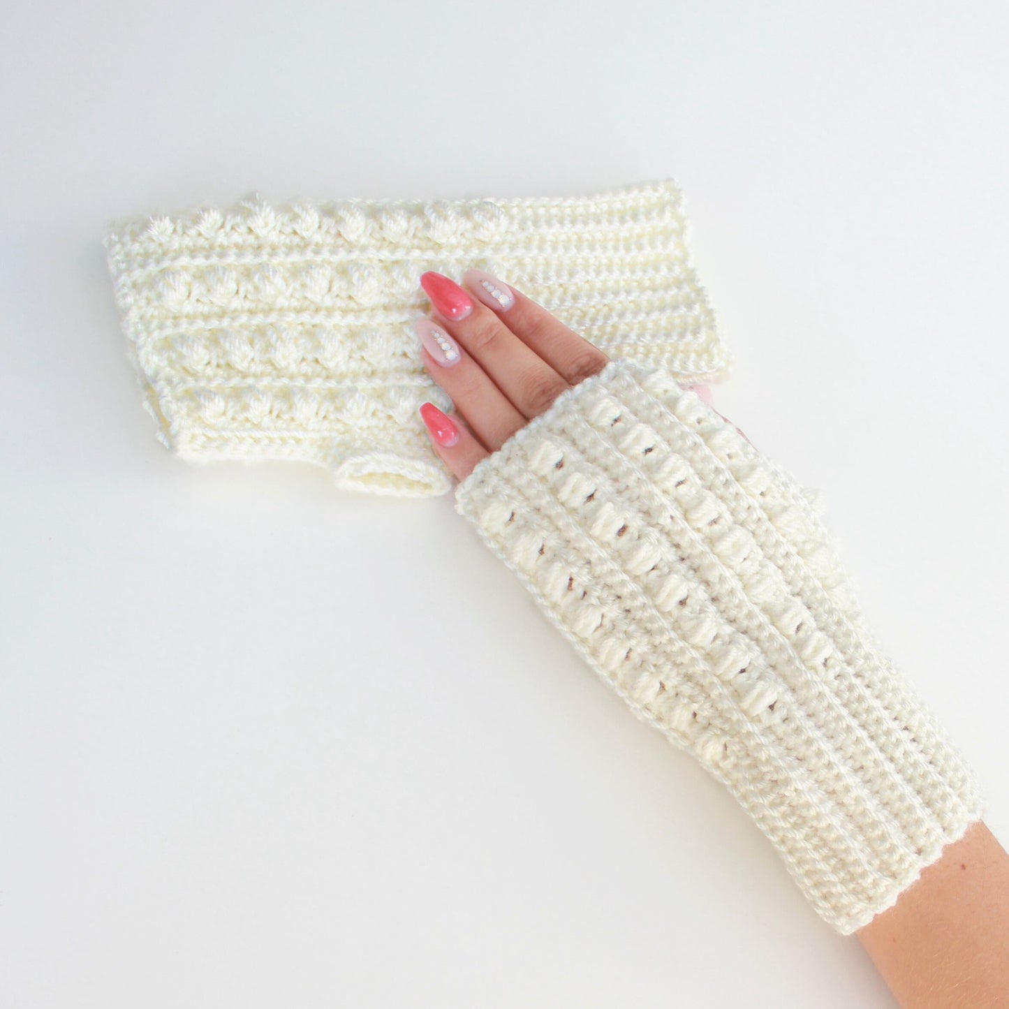 May include: A pair of white crocheted fingerless gloves with a textured design. The gloves are shown on a white background.