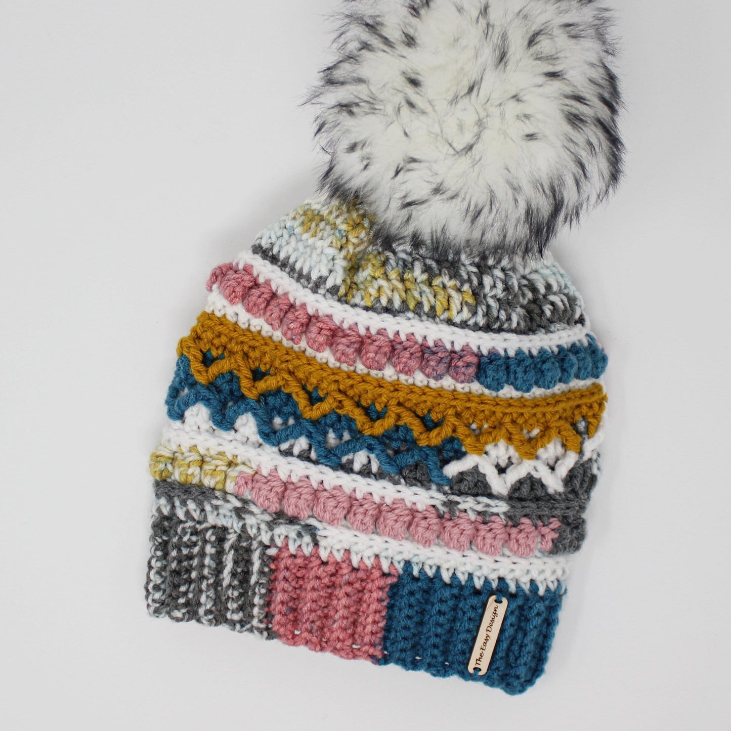May include: A colorful crocheted beanie with a white pom pom on top. The beanie has stripes of gray, white, pink, blue, yellow, and green. The beanie has a label that says "The Easy Designer".