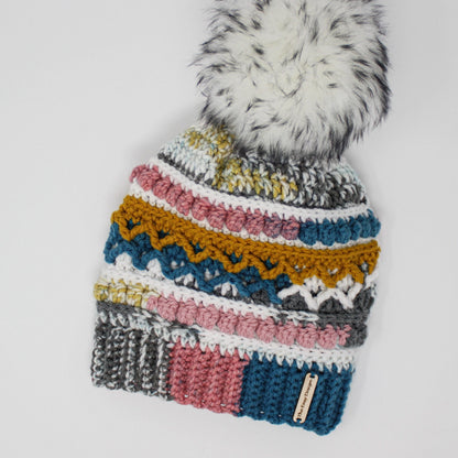 May include: A colorful crocheted beanie with a white pom pom on top. The beanie has stripes of gray, white, pink, blue, yellow, and green. The beanie has a label that says "The Easy Designer".
