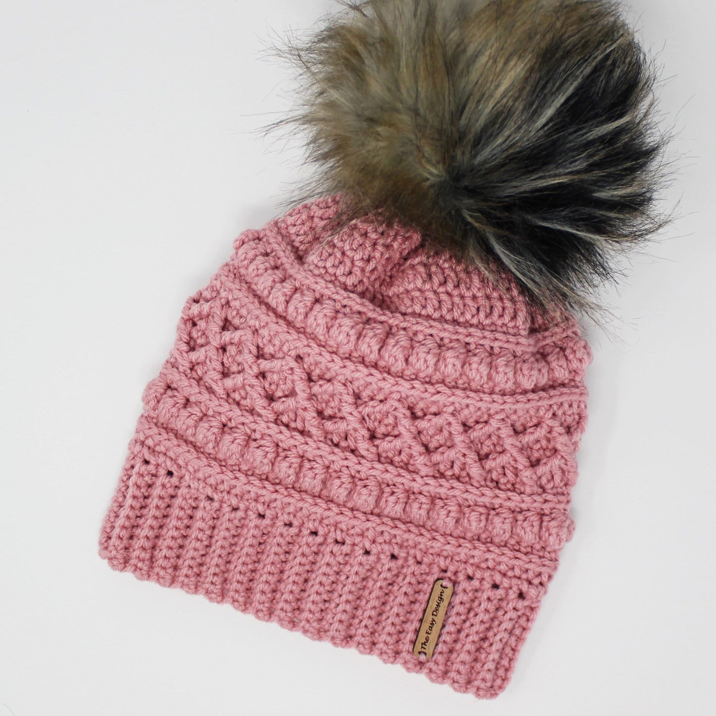 May include: A pink crocheted beanie with a black and brown faux fur pom pom. The beanie has a textured design and a brown leather tag that says "The Cozy Designer".
