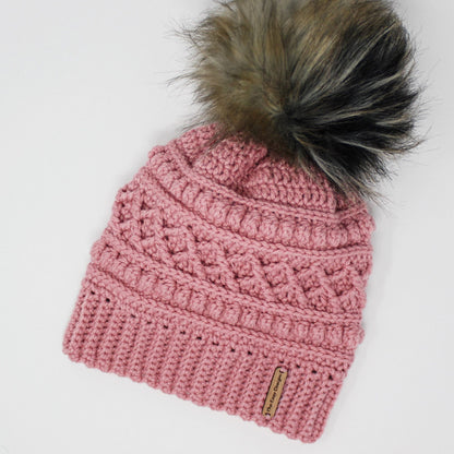 May include: A pink crocheted beanie with a black and brown faux fur pom pom. The beanie has a textured design and a brown leather tag that says "The Cozy Designer".