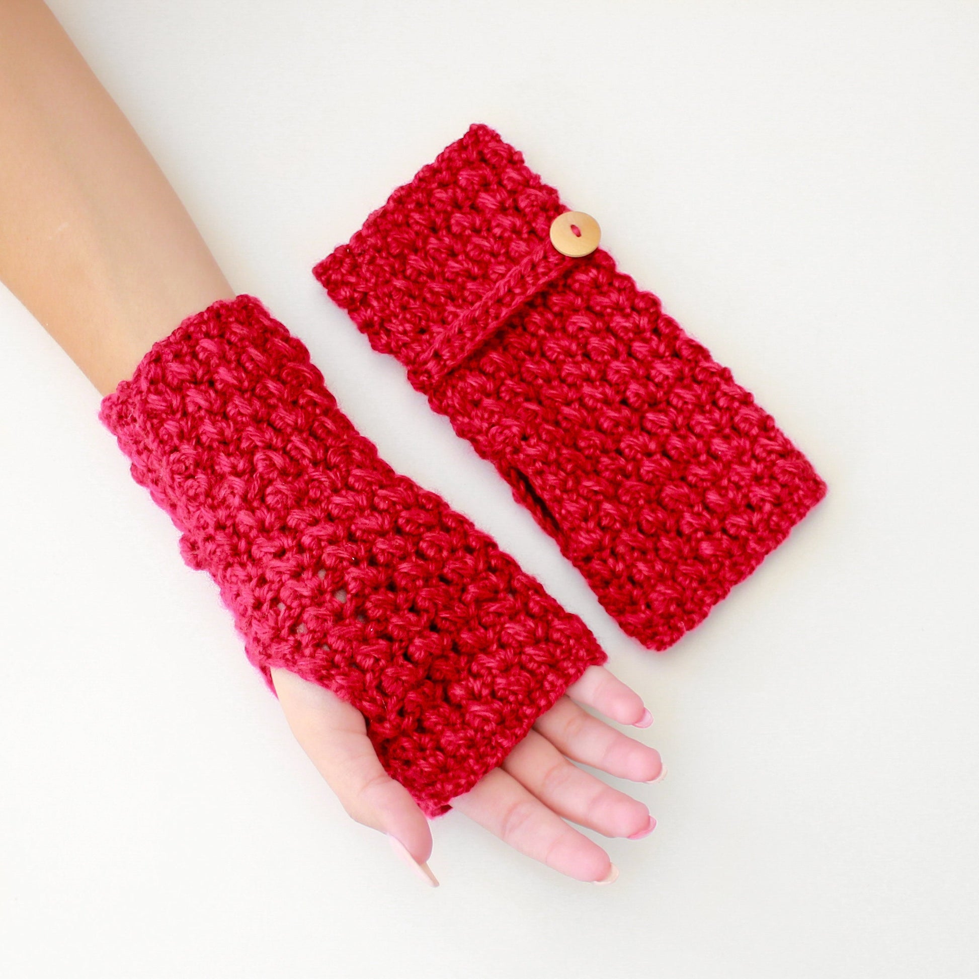 May include: A pair of red crocheted fingerless gloves with a button detail. The gloves are made with a textured stitch and are a warm and cozy accessory.