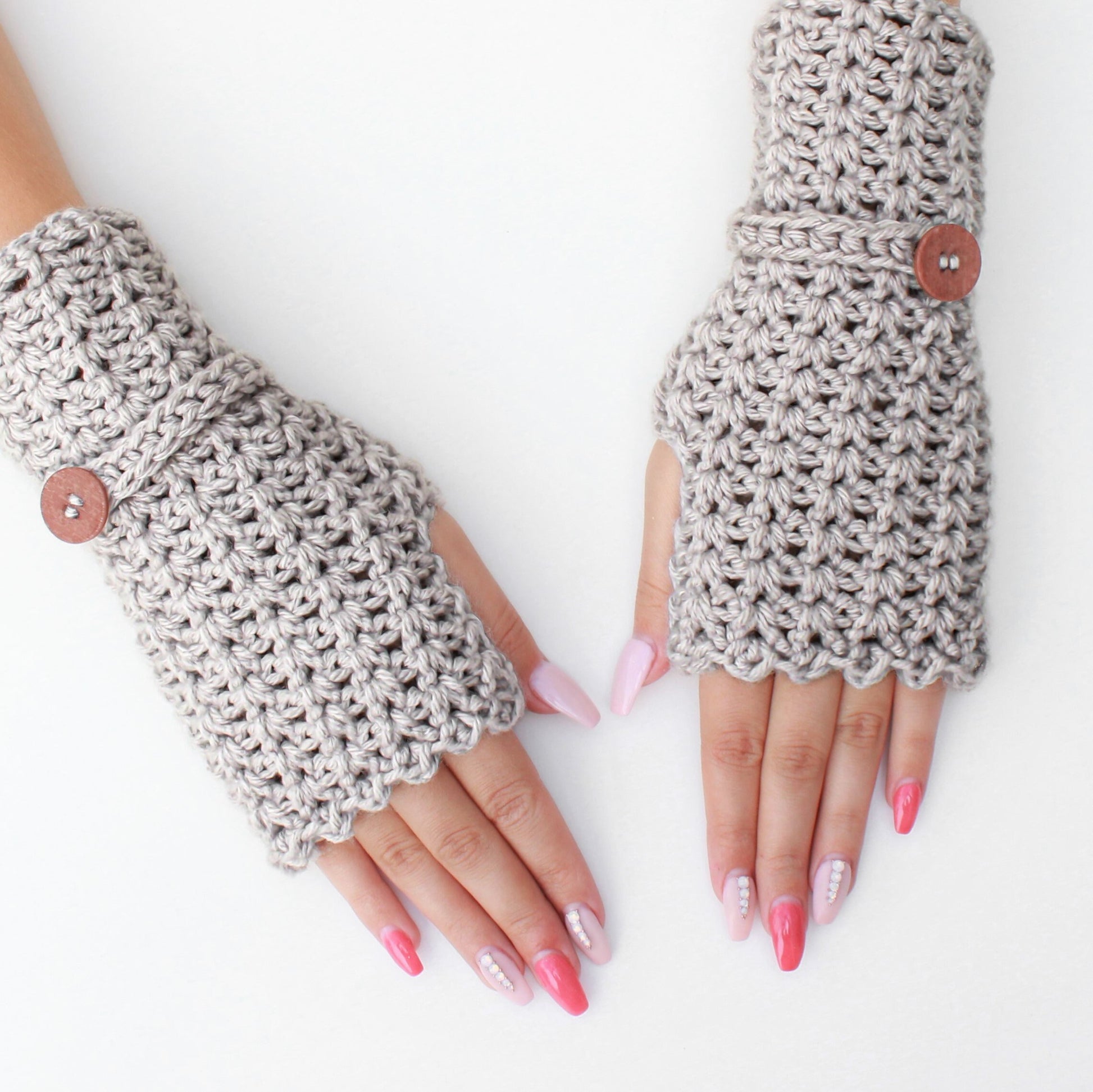 May include: A pair of gray fingerless gloves with a crocheted design and wooden buttons. The gloves are worn on a person's hands with pink nail polish.