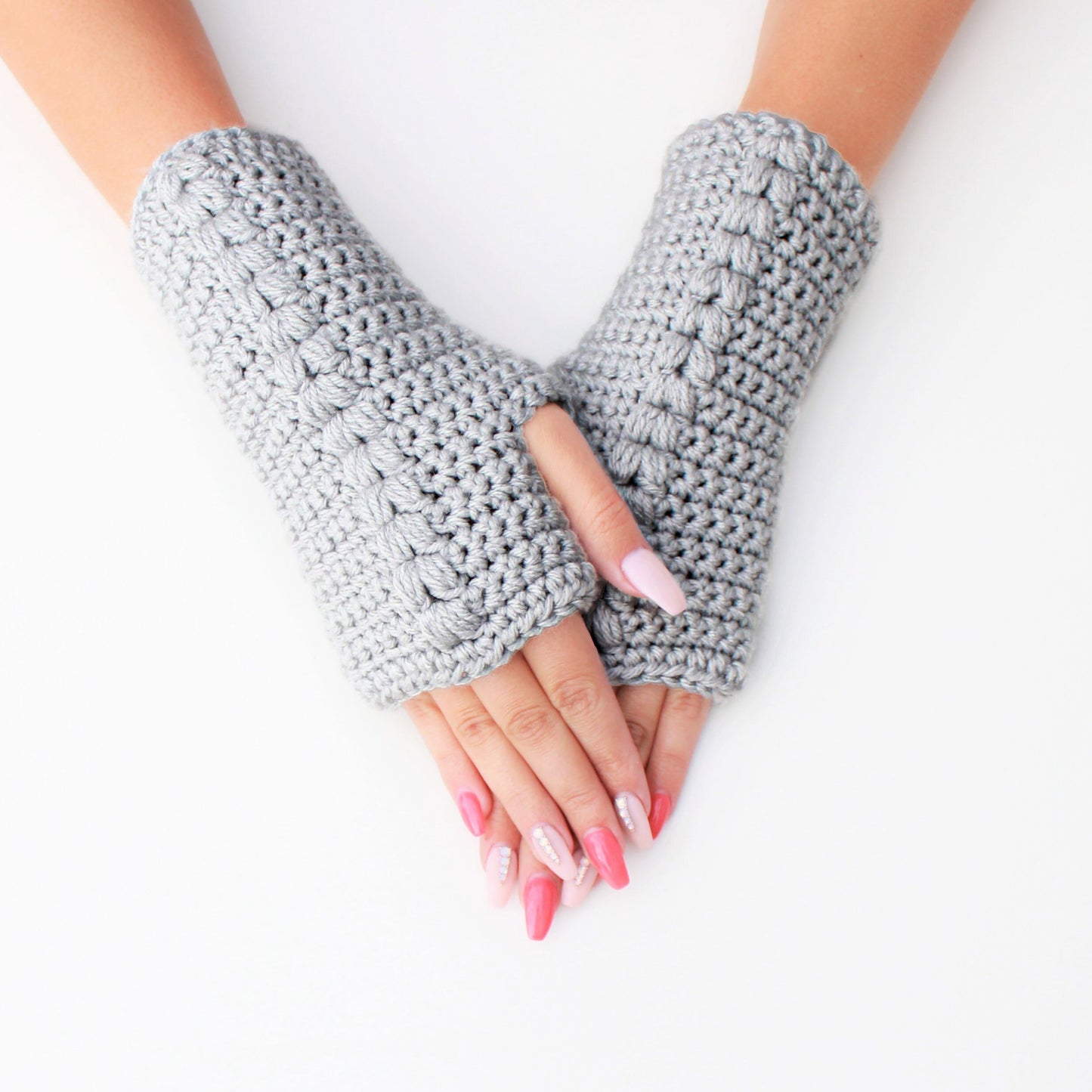 May include: A pair of gray fingerless gloves with a textured knit pattern. The gloves are worn on a person's hands with pink nail polish.