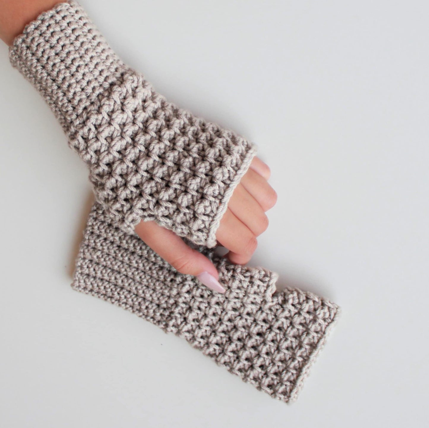 May include: A pair of beige crocheted fingerless gloves. The gloves are made with a textured stitch and have a simple, minimalist design.
