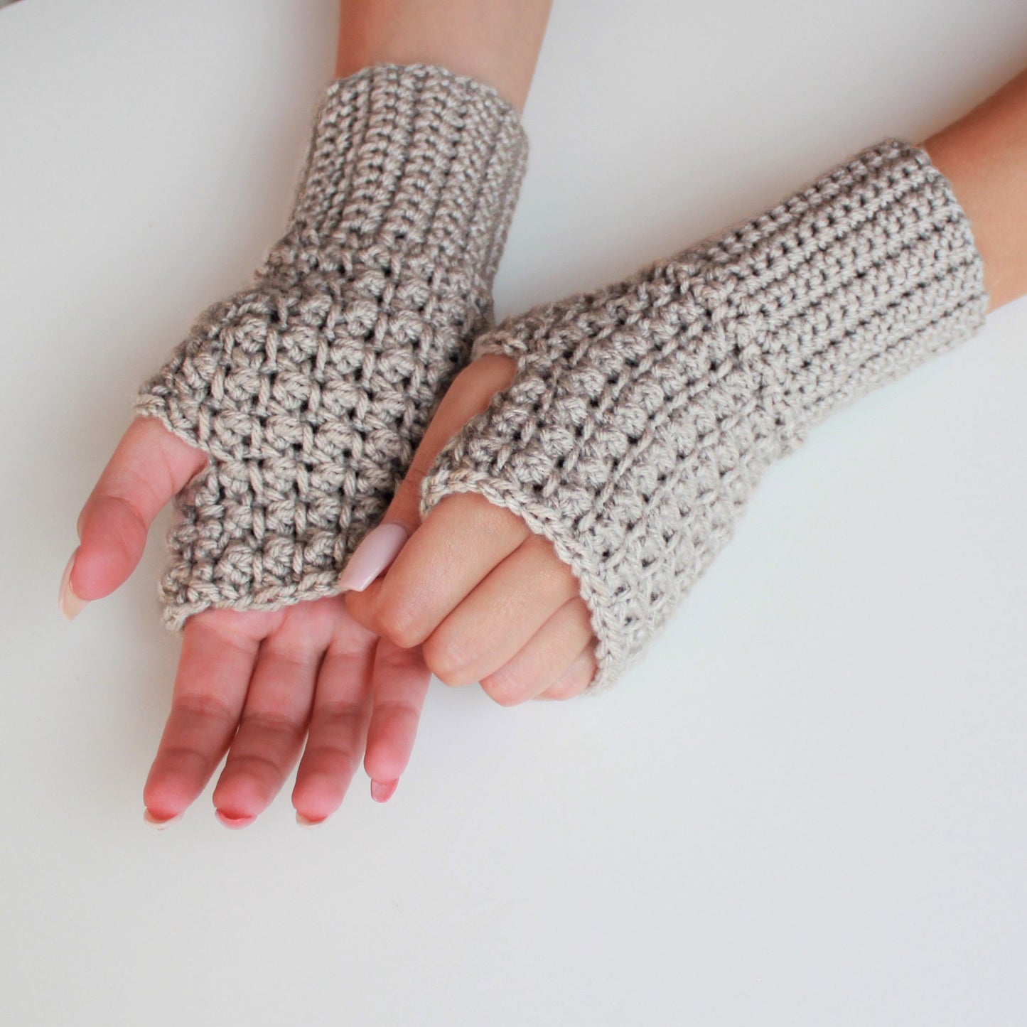 May include: A pair of beige crocheted fingerless gloves. The gloves are made with a textured stitch and have a simple, minimalist design.