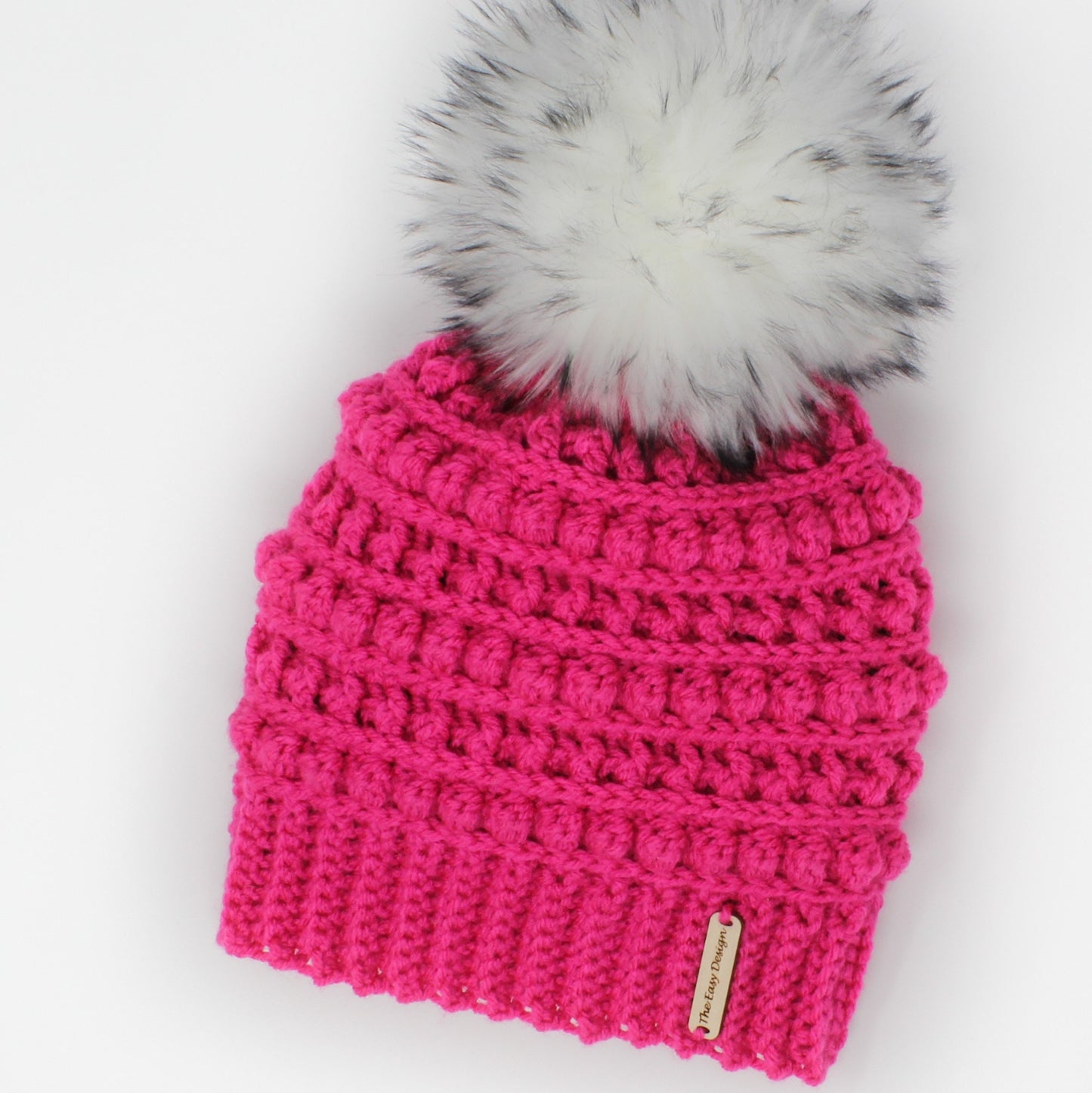 May include: A pink crocheted beanie with a white faux fur pom pom. The beanie has a textured stitch pattern and a small brown tag that says "The Etsy Designer".