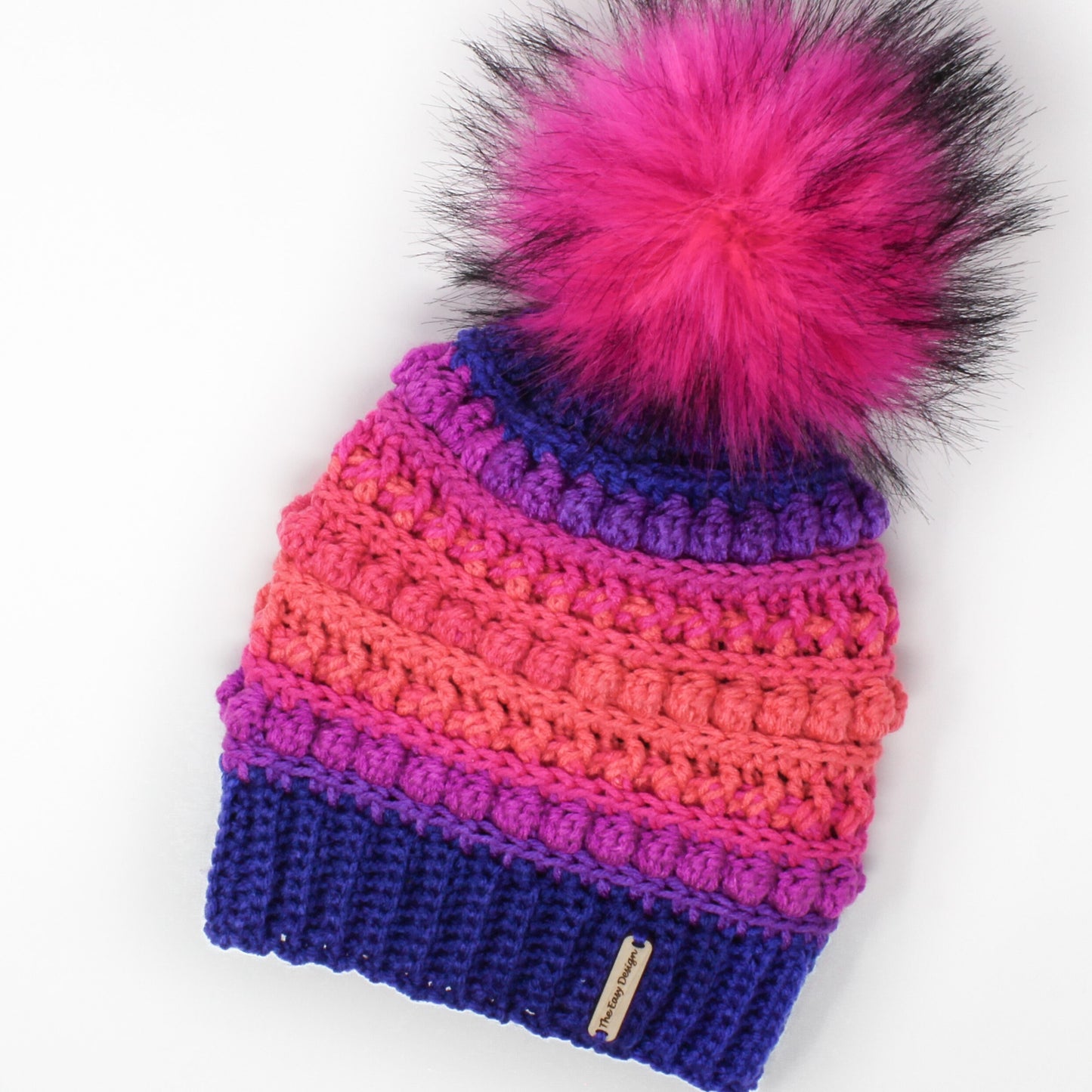 May include: A colorful crocheted beanie with a pink faux fur pom pom. The beanie is made in a gradient of pink, purple, and blue.