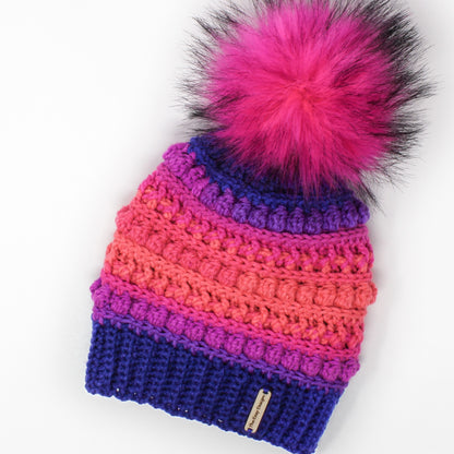 May include: A colorful crocheted beanie with a pink faux fur pom pom. The beanie is made in a gradient of pink, purple, and blue.
