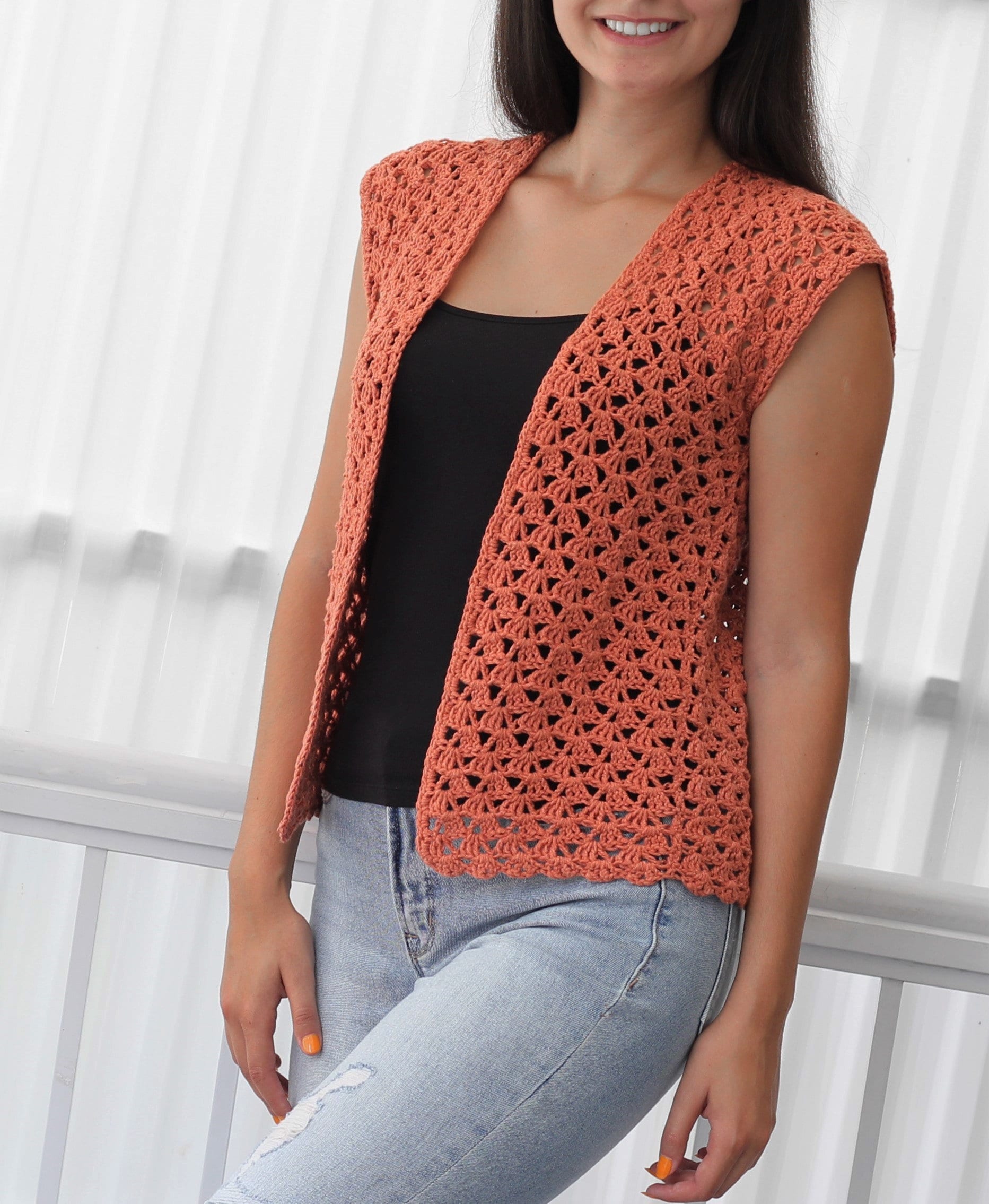 May include: A crocheted sleeveless vest in a terracotta color with an open stitch pattern. The vest is worn over a black tank top and blue jeans.
